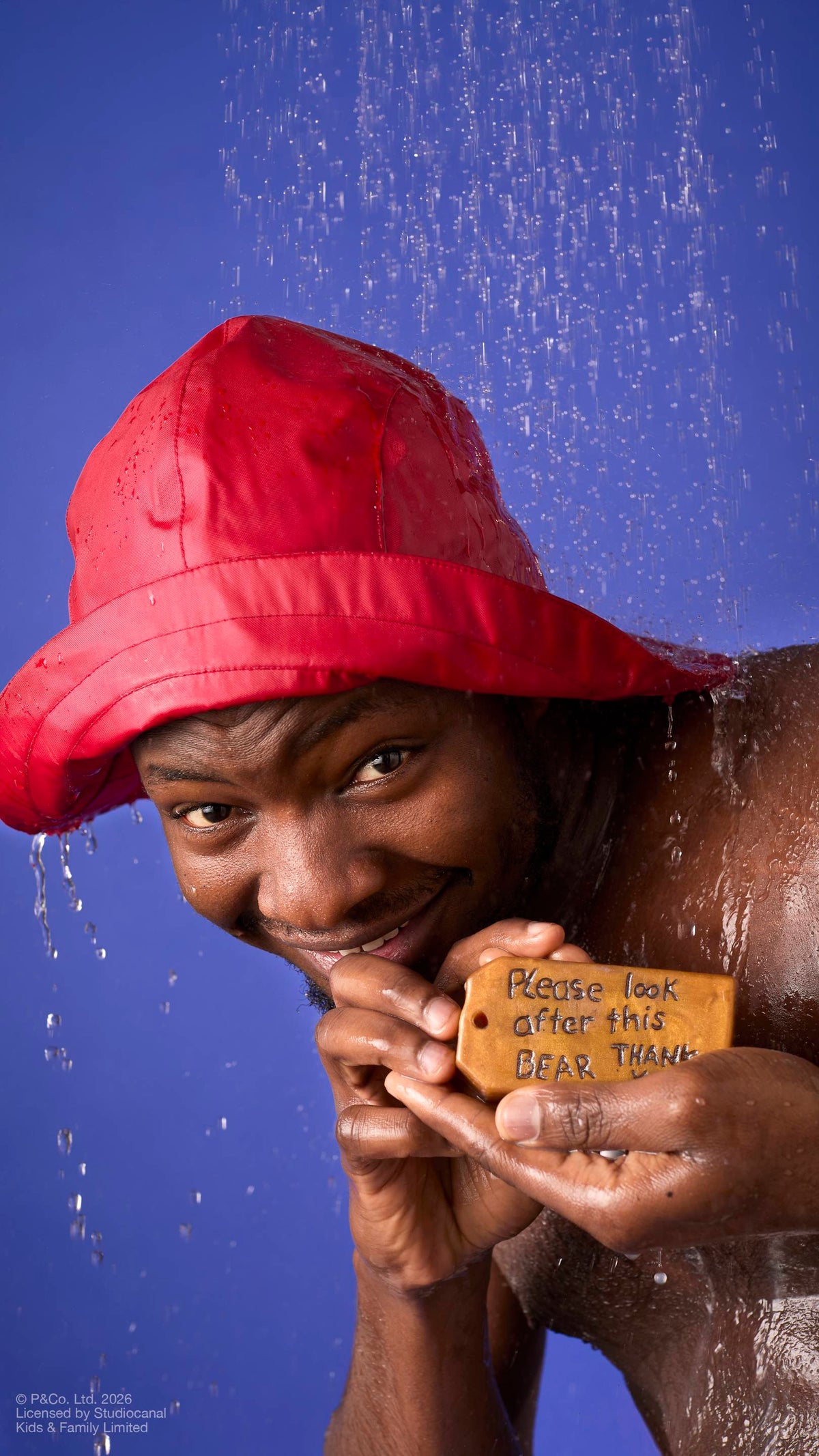 A model wearing a red rain hat stands under running shower water. They hold a brown, luggage-tag-shaped soap featuring the embossed message ‘Please look after this bear, thank you’. Droplets cascade down against a solid blue background.